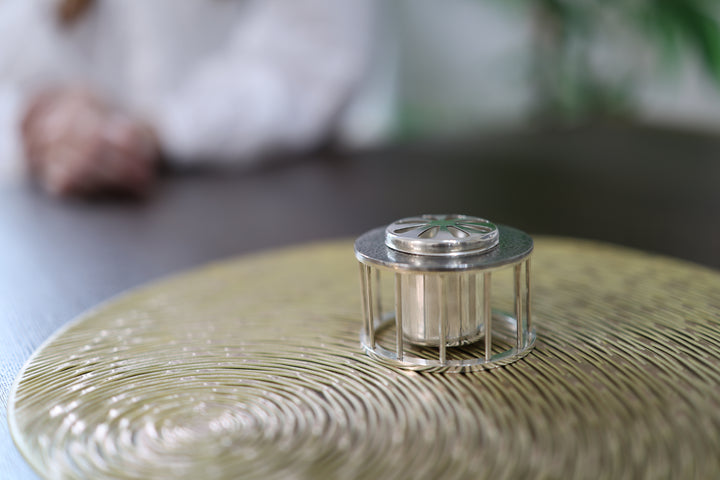 Pure silver incense burner on a textured metallic table with a seated person softly blurred behind it.