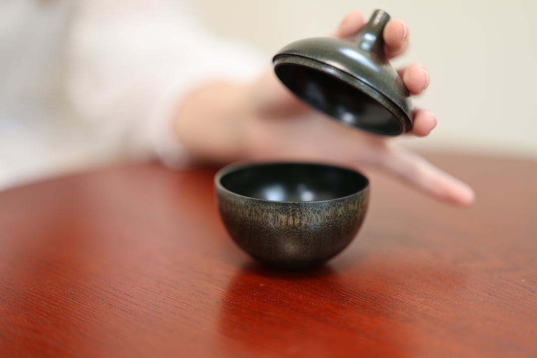 A hand lifting the stemmed lid from Bulb: Sesame on a lacquered table.