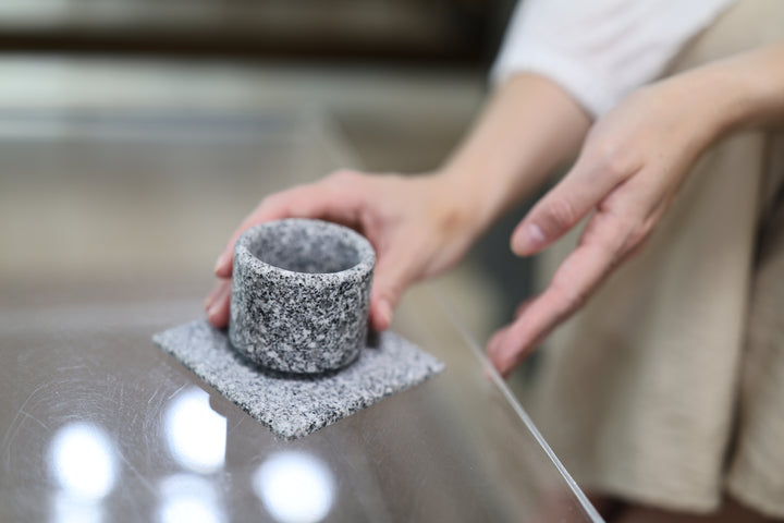 A cylindrical stone cup on the Makabe Stone Art Plate on a reflective glass tabletop, with hands nearby.