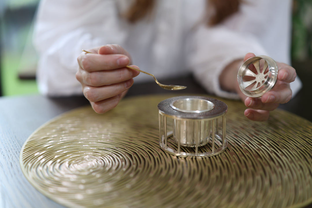 Person lifting the vented lid beside the open pure silver incense burner on a textured table.
