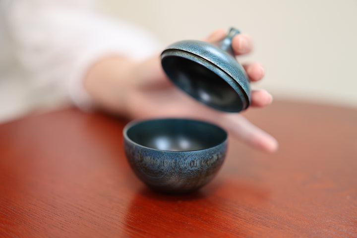 A hand lifting the tapered lid above the rounded lower body of Bulb: Hyacinth on a red table.