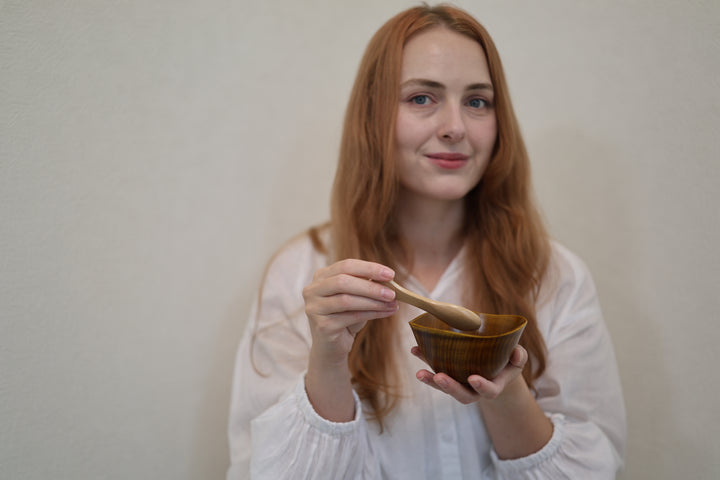 Adult model holding the RIN Bowl M Mimosa and a wooden spoon at chest height against a pale wall.