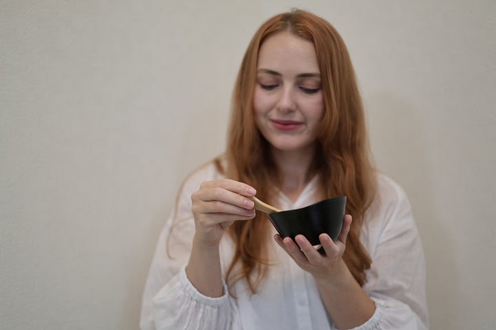 Adult model holding the RIN Bowl M Sesame and a wooden spoon at chest height against a pale wall.