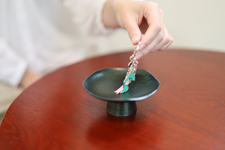 A hand lifts keys from the shallow blue-green lacquered surface of Floating Leaf Hyacinth.