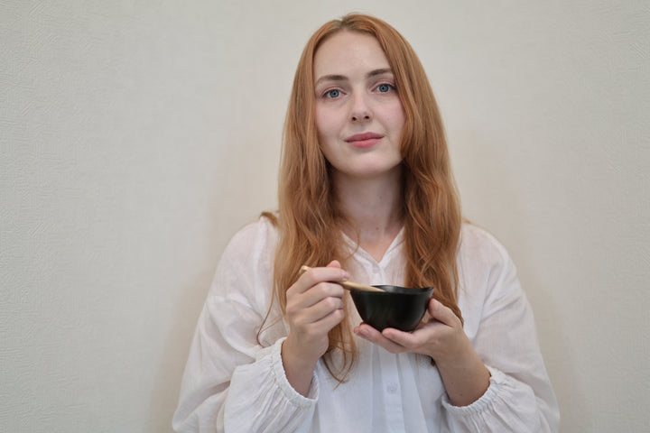 Adult model holding the RIN Bowl S Sesame and a wooden spoon at chest height against a pale wall.