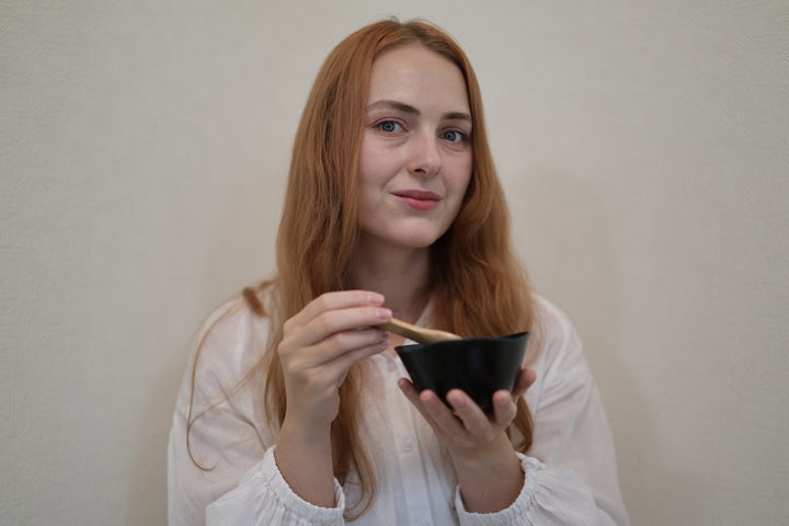 Seated adult model holding the RIN Bowl M Hyacinth and a wooden spoon against a pale wall.