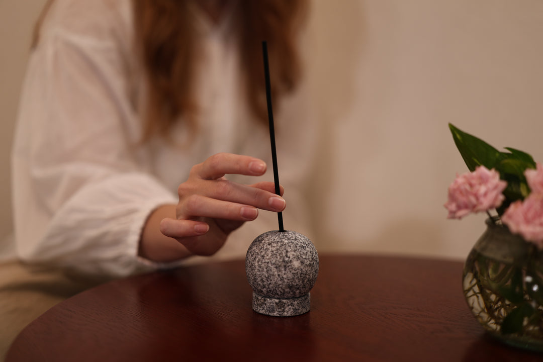 A hand placing an unlit black incense stick in the Makabe Stone Incense Stone on a round wooden table beside pink flowers.