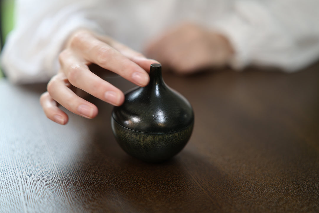 A hand approaching the narrow stem of Bulb: Sesame on a dark tabletop.