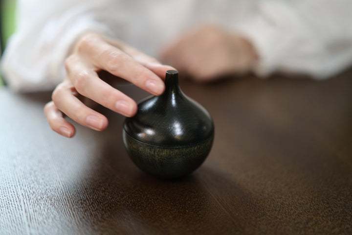 A hand approaching the narrow stem of Bulb: Sesame on a dark tabletop.