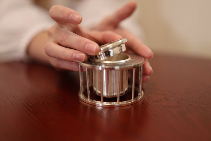 Pure silver incense burner on a warm wooden table beside flowers, with a hand touching the lid.