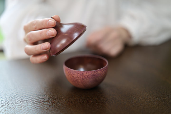 A hand holding the compact lilac and wooden lidded object above a dark table