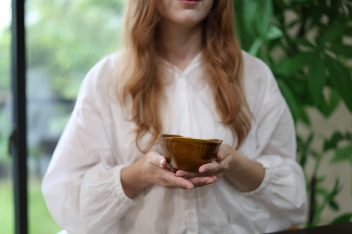 Adult model supporting the empty RIN Bowl M Mimosa in both hands near a window with greenery behind.