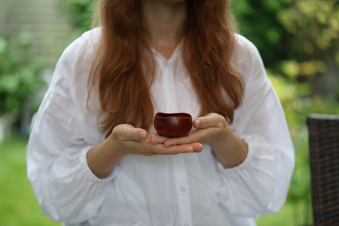 A person in a white blouse presenting the berry-red lacquerware vessel in both hands with greenery behind.