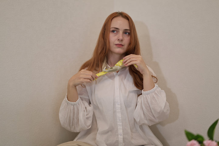 A wearer adjusting the narrow patterned silk scarf at the neck against a plain wall.