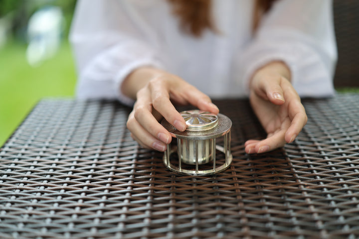 Pure silver incense burner on a woven outdoor table, with a person reaching toward the lid.