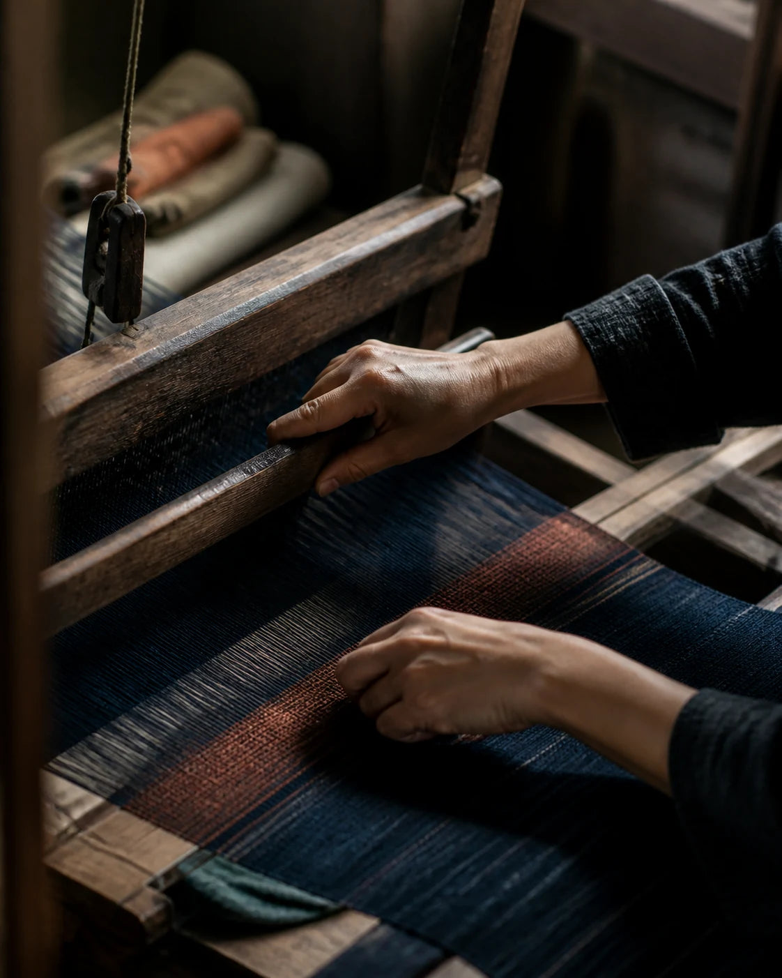 Anonymous artisan hands weaving indigo and muted rust textile on a wooden handloom